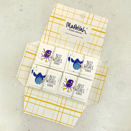 An open Mapayah KIT Strangelings stationery folder with a yellow grid pattern, showing the Mapayah logo, a personalised notepaper sheet with the name Kiara, and two bundles of cards tied with string — one printed with a purple alien on ice skates and one with a blue penguin — set against a solid light beige background.