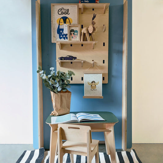 A birch plywood NOTCH wall shelving system mounted on a dark teal display wall above a children's wooden desk and chair, displaying framed prints, a woven wooden reindeer figure, a toy car, and a hanging "Stay Blessed" print, with a paper-wrapped plant on the desk.