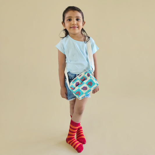 A young girl stands against a solid beige background wearing a light blue t-shirt, denim shorts, red and orange striped socks, and holding a small quilted crossbody bag with a bug and checkered pattern.