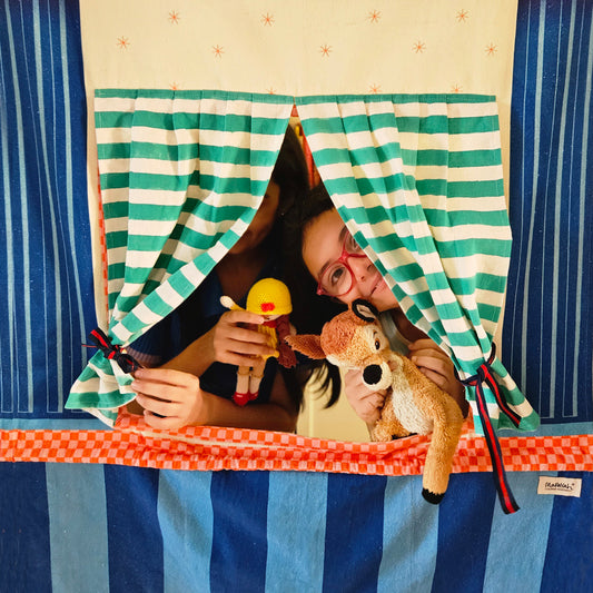 A child peeks through the green and white striped stage curtains of the Mapayah STAGE Theatre doorway curtain, holding a crocheted doll puppet and a plush deer, with the orange checkered trim visible below.