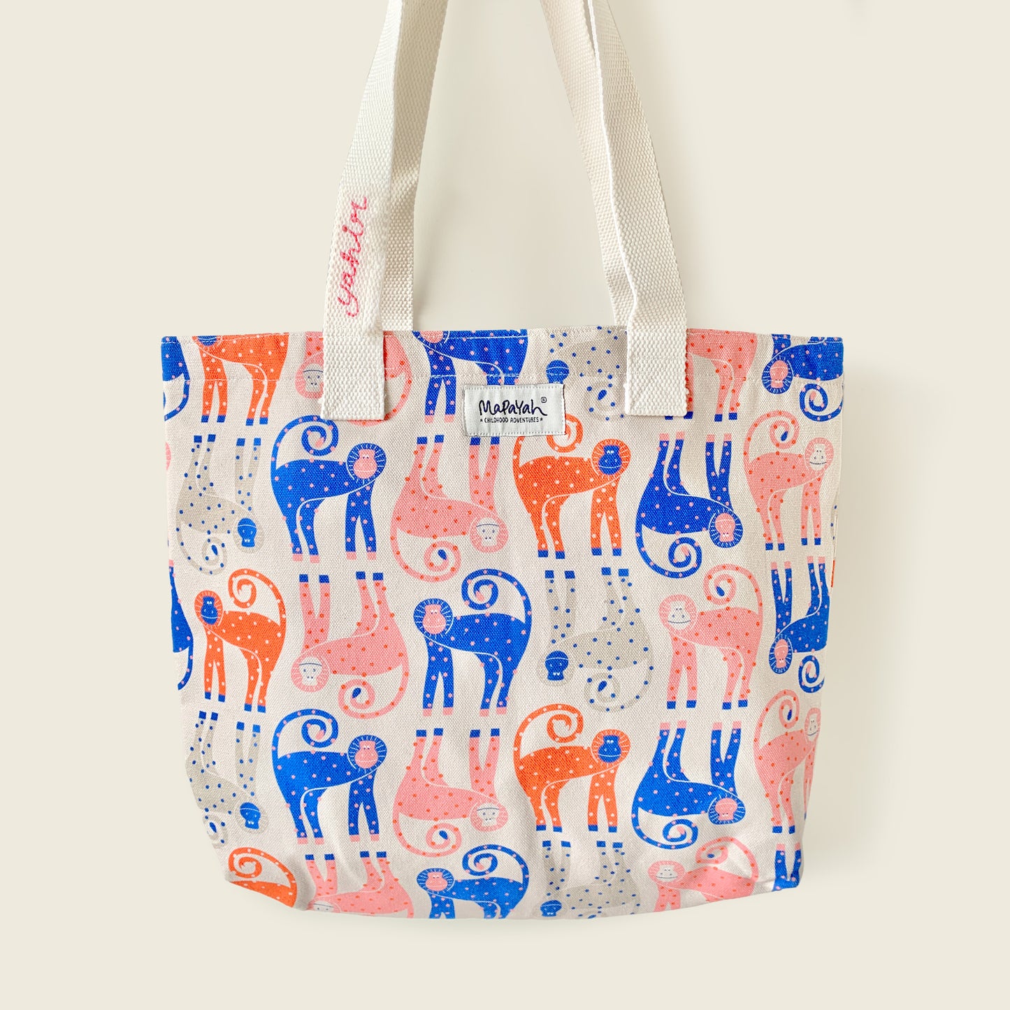 A canvas tote bag with a repeating pattern of stylized monkey figures in blue, pink, and orange on a cream background, featuring customised name on white shoulder straps and a small 'Mapayah' brand label.
