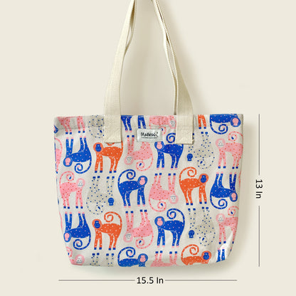 A tote bag with a colorful pattern of stylized monkey figures in blue, pink, and orange on a off white background, with off white shoulder straps. Measurement lines indicate dimensions of 15.5 inches width and 13 inches height.