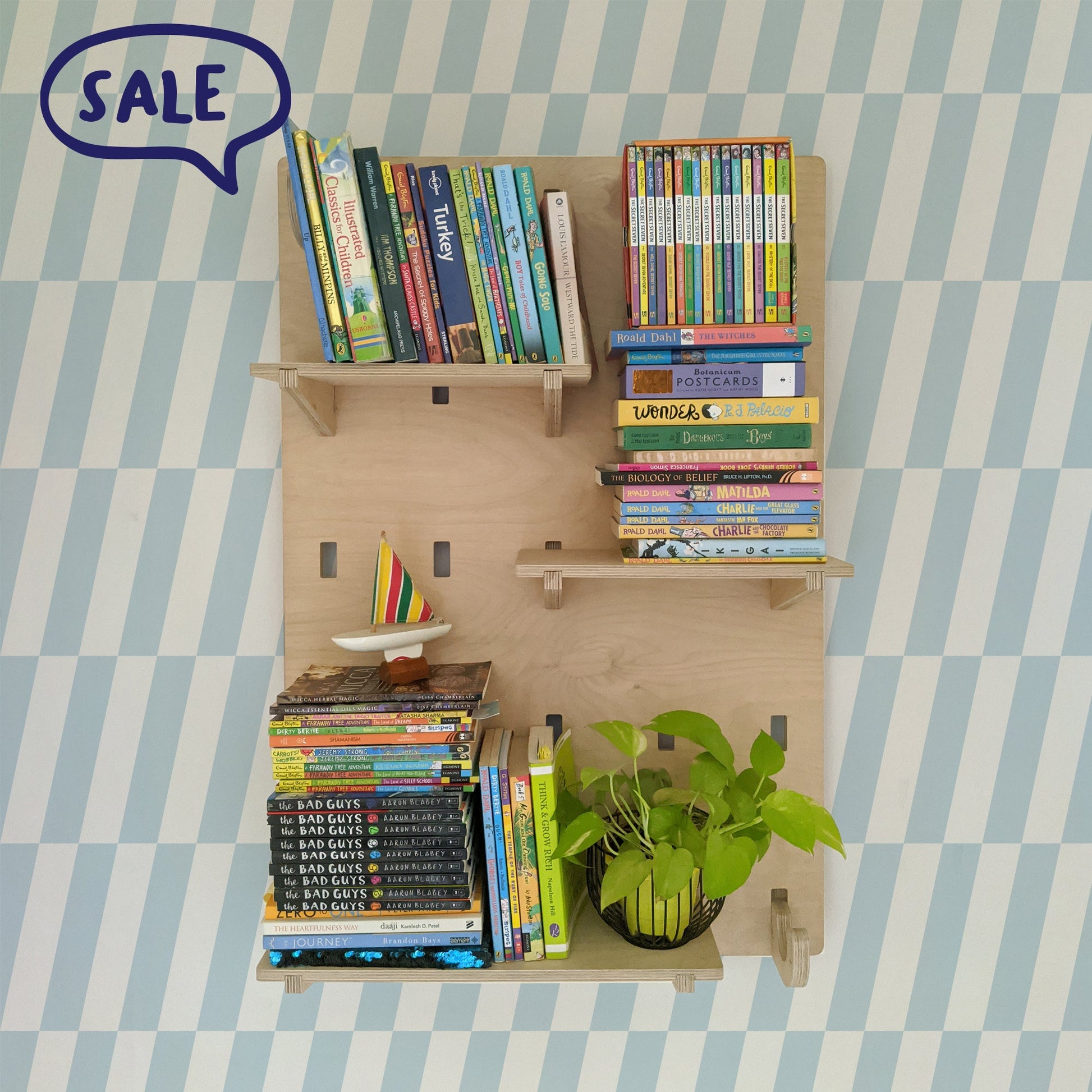 A birch plywood NOTCH wall shelving system mounted on a blue and white checkered wallpaper wall, holding stacks of children's books, a toy sailboat, and a potted plant.