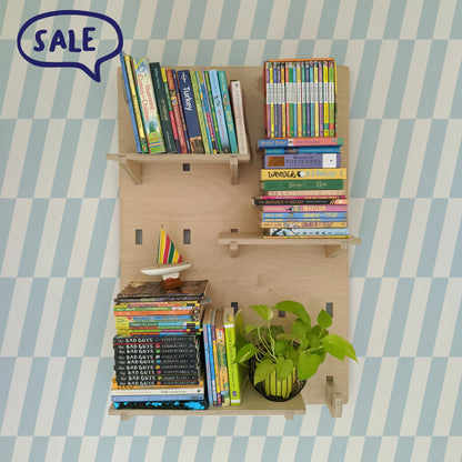 A birch plywood NOTCH wall shelving system mounted on a blue and white checkered wallpaper wall, holding stacks of children's books, a toy sailboat, and a potted plant.