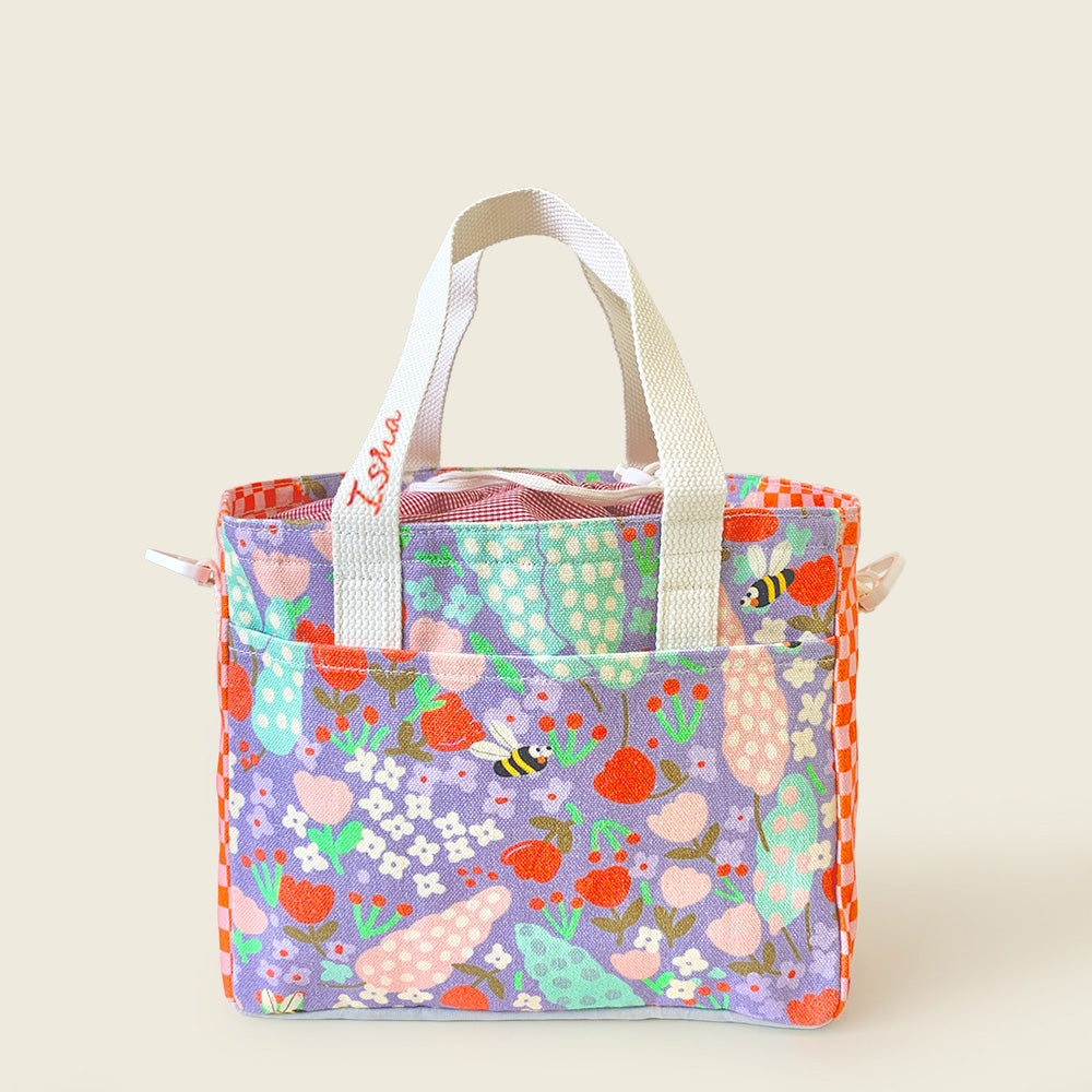 A fabric lunch bag with a colorful floral and bee print on a lavender background, featuring off white handles, set against a solid light beige background.