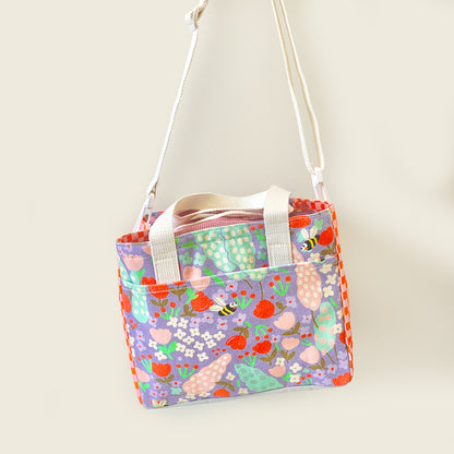 A small lunch bag with a colorful spring floral and bee print, featuring a white shoulder strap and top handles, shown against a plain light beige background.