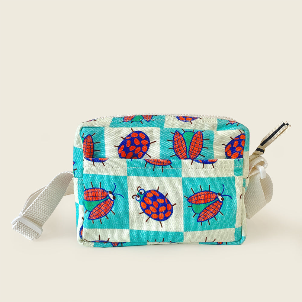 A small canvas pouch with a white and turquoise checkered pattern and red ladybug illustrations, featuring a white shoulder strap and a silver zipper pull, set against a solid light beige background.