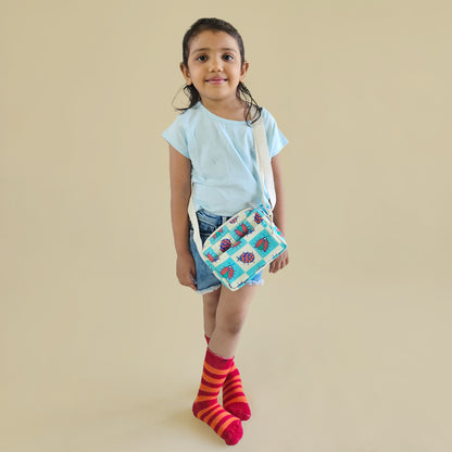 A young girl stands against a solid beige background wearing a light blue t-shirt, denim shorts, red and orange striped socks, and holding a small quilted crossbody bag with a bug and checkered pattern.