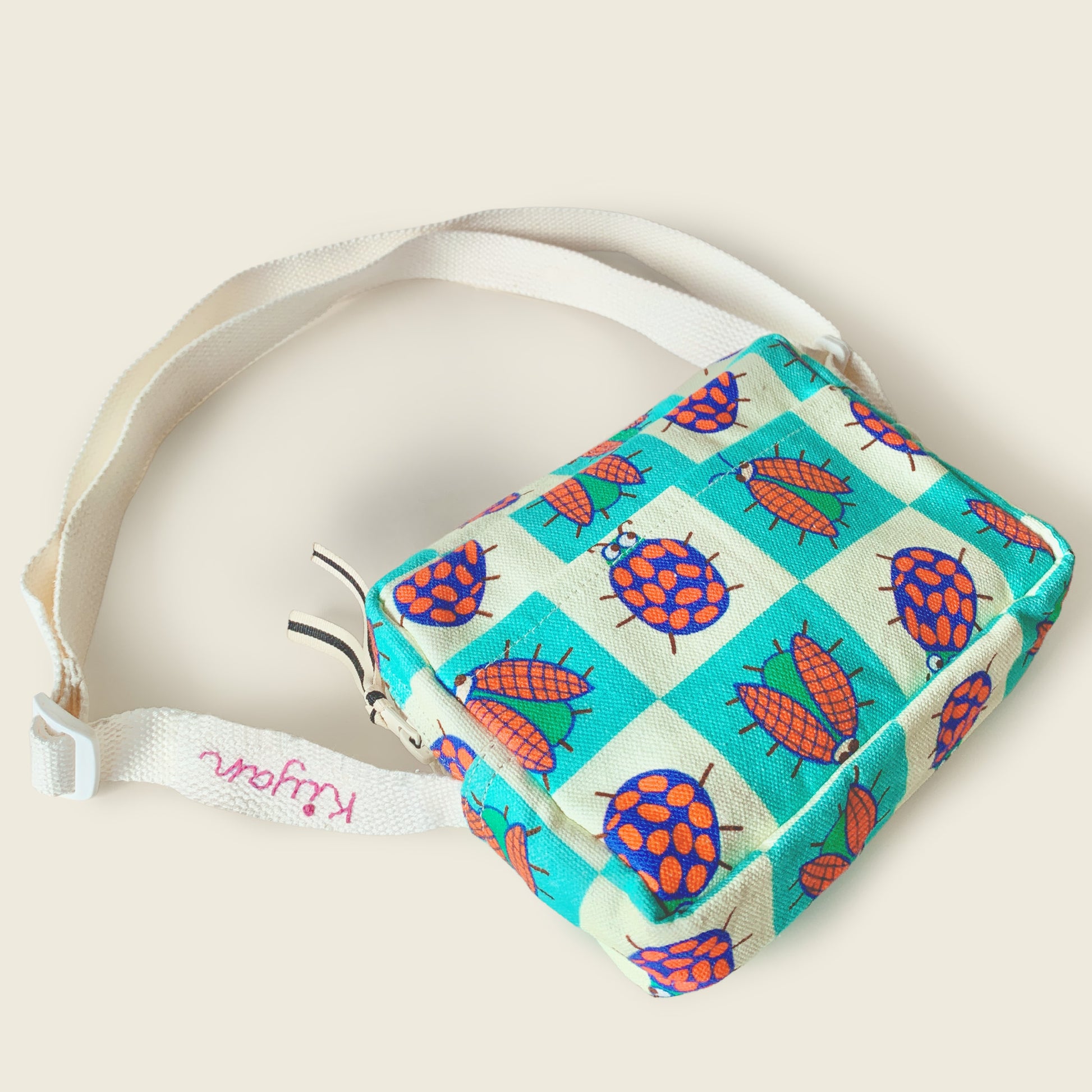 A small fabric shoulder bag with a colorful ladybug print on a teal and cream checkered background, featuring an adjustable cream canvas strap with a pink embroidered customised name.