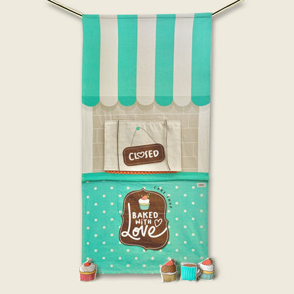 A teal and cream striped awning doorway curtain with a checkered orange serving window, a "CLOSED" sign hanging inside the window, and a teal polka-dot counter printed with a "Baked With Love" cake shop logo, with stuffed cupcake and mug props at the base, set against a solid cream background.