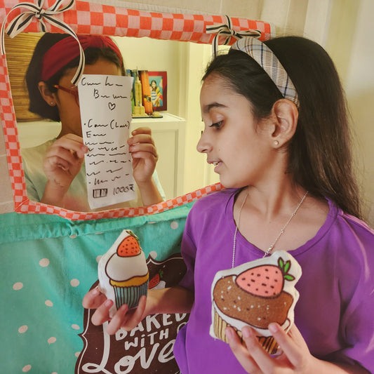 A young girl in a purple t-shirt stands at the serving window of the Mapayah Stage Café doorway curtain, holding a stuffed cupcake and reading a handwritten order receipt, with another child visible through the window taking the order.