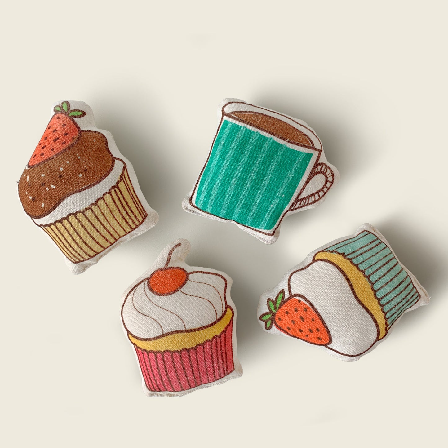 Four soft stuffed play props included with the Mapayah Stage Café: a chocolate cupcake topped with a strawberry, a teal coffee mug, a pink cupcake topped with a cherry, and a strawberry, laid out on a solid cream background.