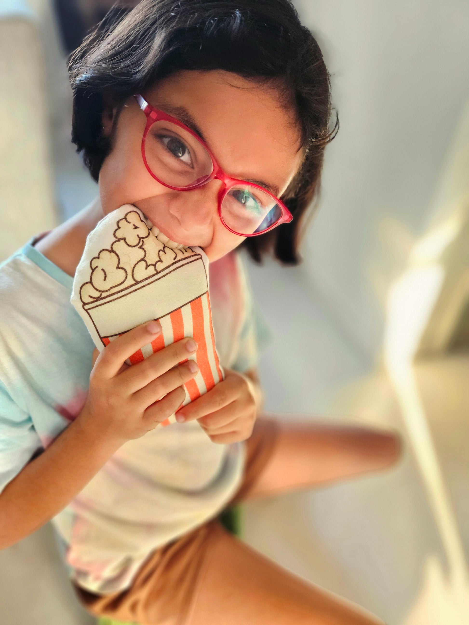 A young girl wearing red glasses holds a stuffed popcorn bucket prop up to her cheek, smiling playfully.