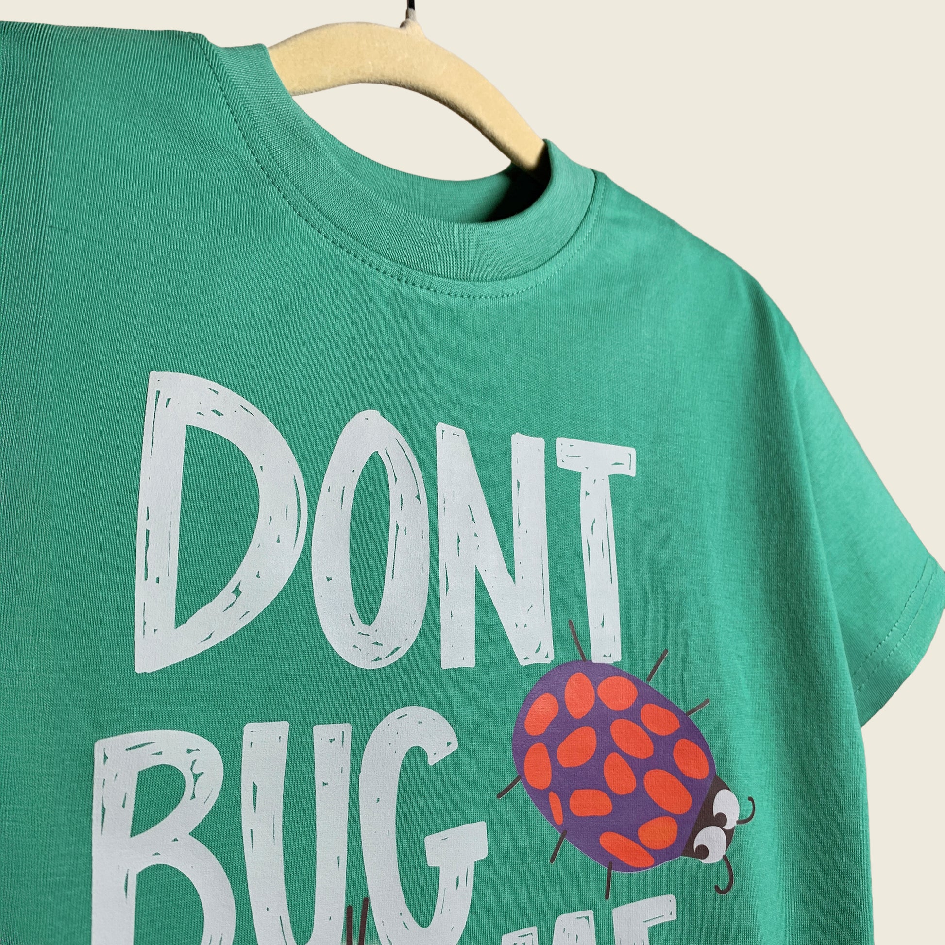 Close-up of green t-shirt with 'Don't Bug Me' text and ladybug graphic on a beige background