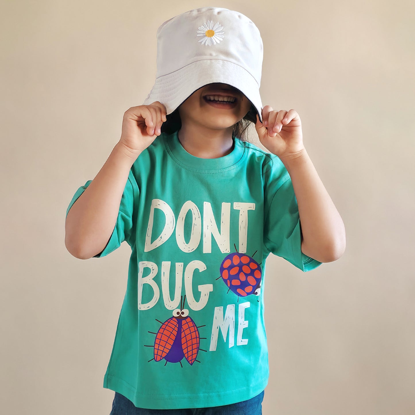 Child wearing a green t-shirt with 'Don't Bug Me' text and a white bucket hat with a flower design.
